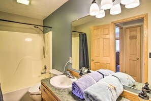 Ein Badezimmer mit Badewanne, Toilette, Waschbecken und einem großen Spiegel. Auf der Ablage liegen Handtücher und im Hintergrund ist eine Holztür zu sehen.