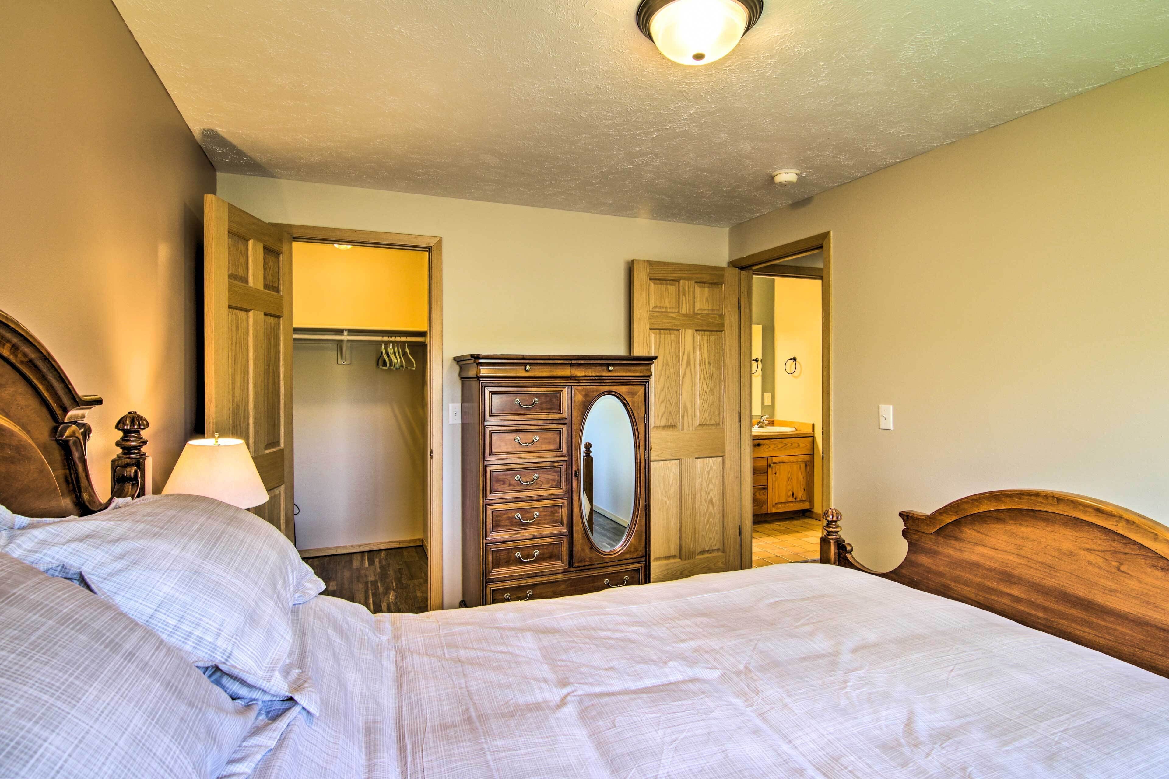 Quarto com cama grande, cómoda de madeira com gavetas, espelho e guarda-roupa com roupas penduradas.