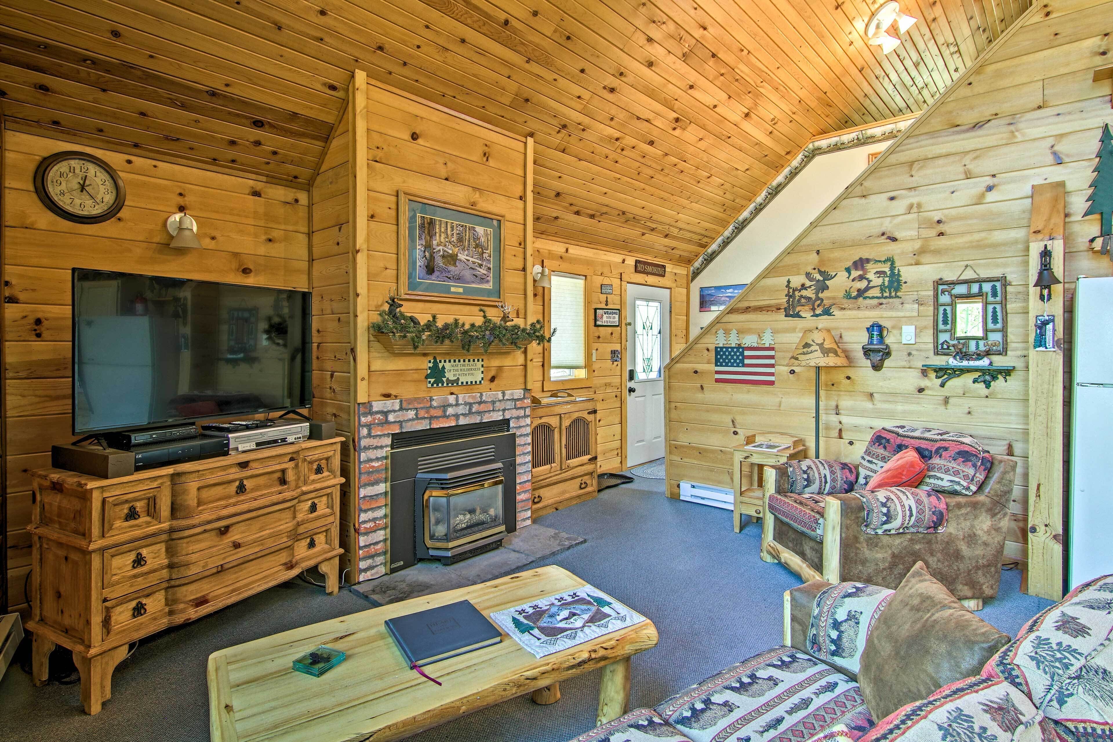 L’intérieur chaleureux d’un chalet avec une cheminée, des meubles en bois, un téléviseur et un escalier menant à un autre niveau.