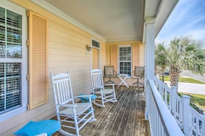 A porch with white rocking chairs and a table, overlooking a street with palm trees.