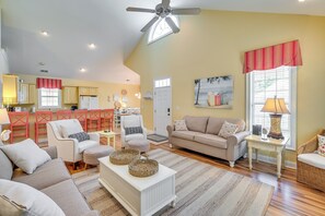 A spacious living room with a high ceiling fan, a dining area with red chairs, and a kitchen with yellow cabinets.