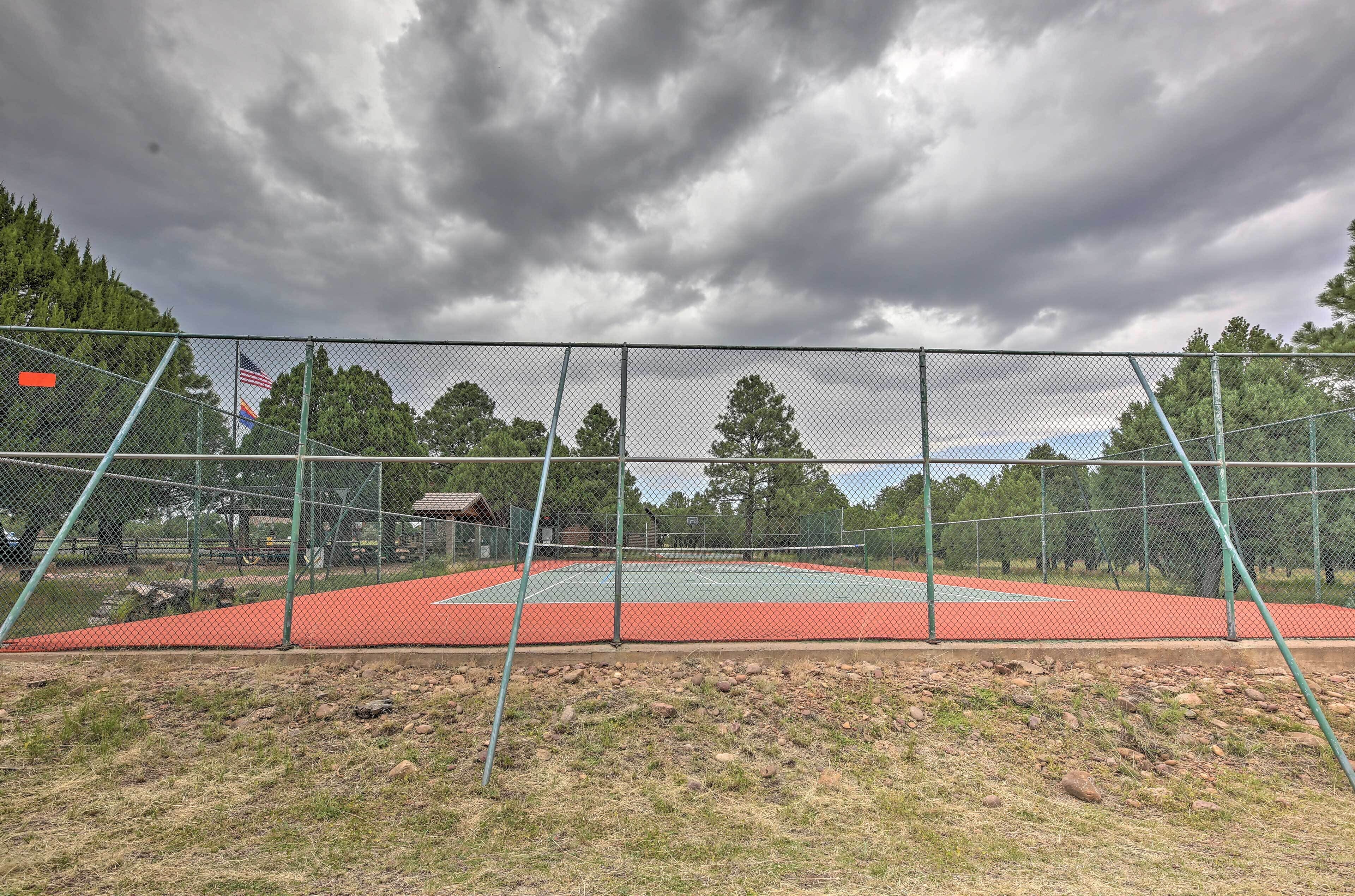 Una cancha de tenis exterior con valla verde, rodeada de árboles y un cielo nublado.