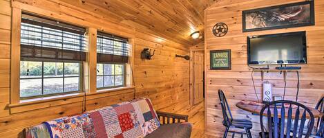 L’intérieur d’un chalet accueillant, avec un plancher en bois, un canapé recouvert d’une courtepointe à motifs, une table à manger avec des chaises, un téléviseur fixé au mur et une fenêtre avec des stores.