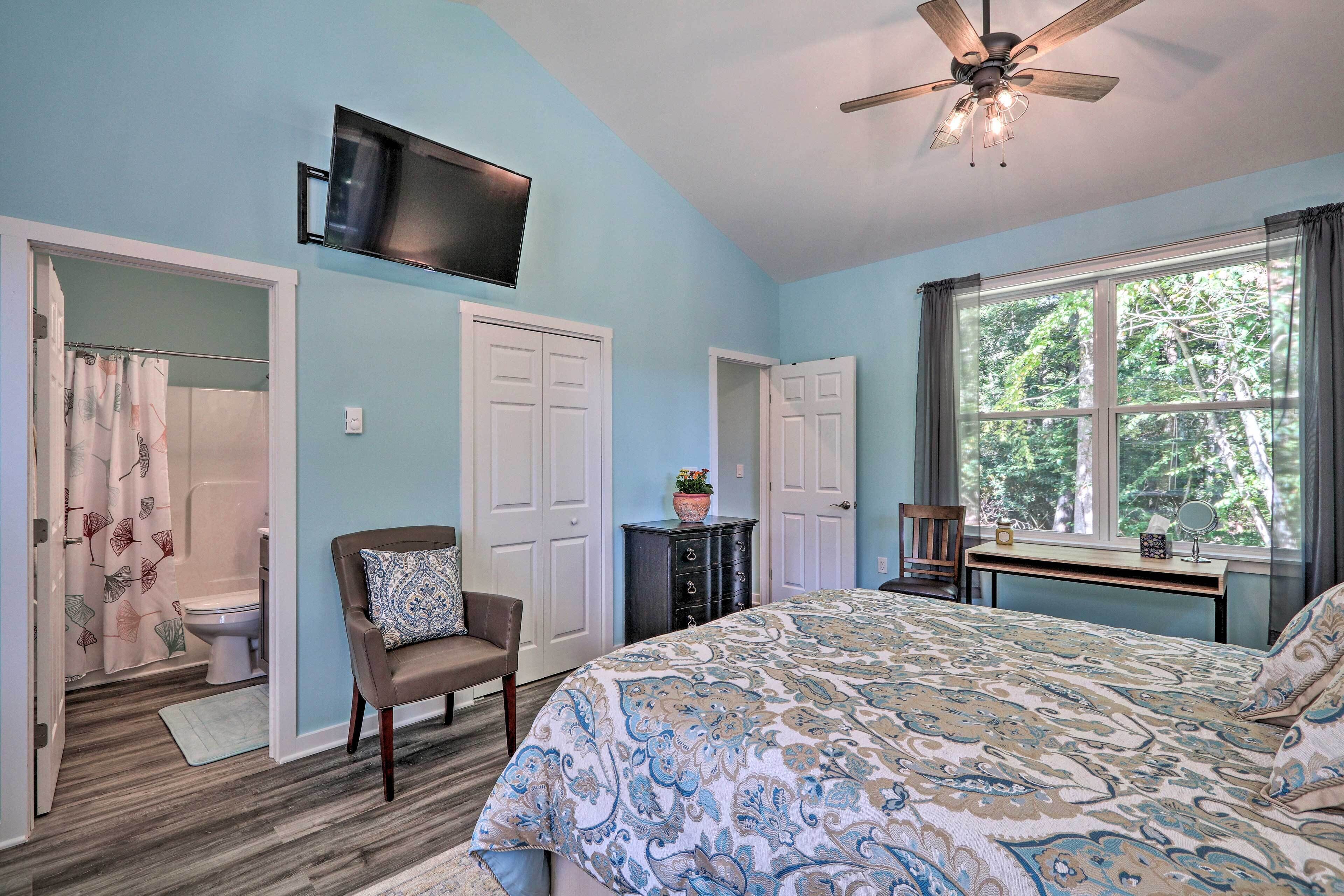 A bedroom with a bed, a TV mounted on the wall, a desk with a chair, and a window with a view of trees.