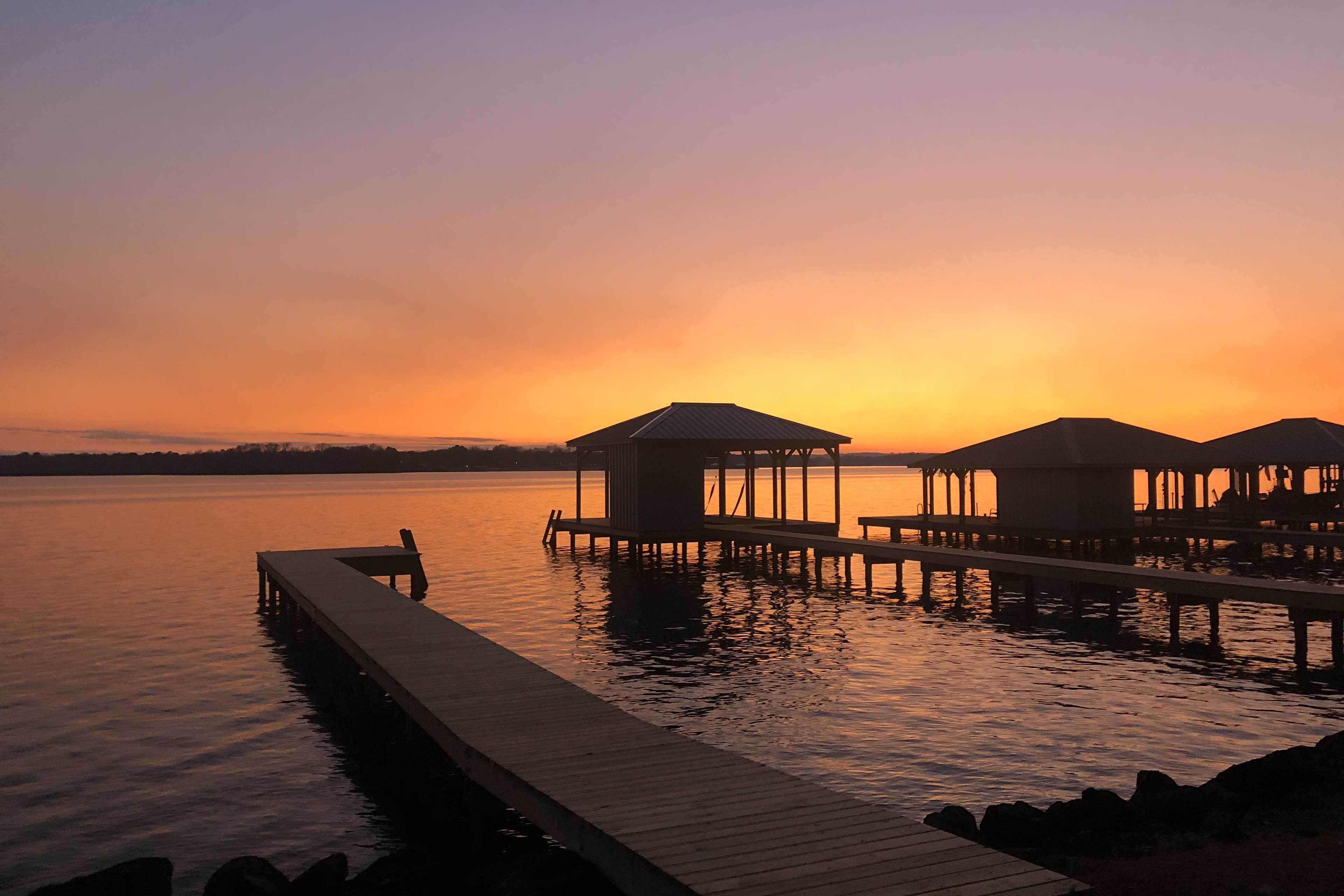 Pôr do sol sobre um lago calmo com cais de madeira e bangalôs sobre a água.