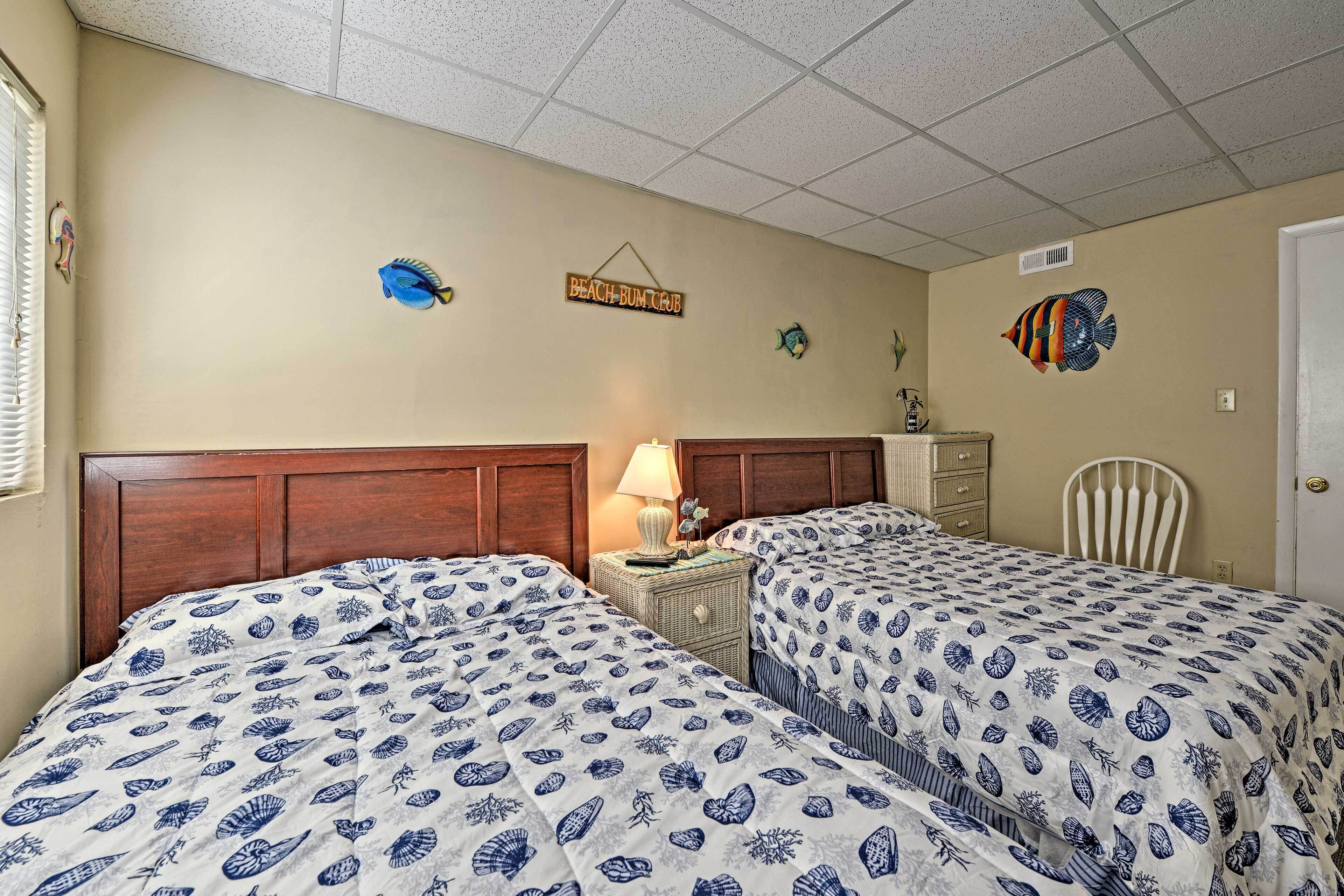 A bedroom with two beds, a nightstand with a lamp, a dresser, and wall decorations including fish and a sign that reads "BEACH BUM BEACH".