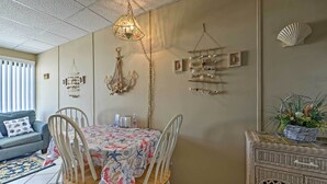 A dining area with a table covered in a patterned tablecloth, two white chairs, a hanging light fixture, wall decorations including a shell and a hanging ornament, and a wicker side table with a plant. - Condo w/ Pool Access on Wildwood Crest Beach! (Wildwood Crest)