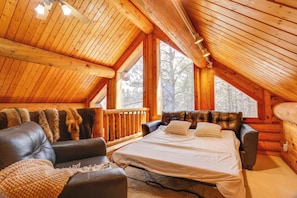 A log cabin bedroom with a large bed, leather sofa, and a view of trees through the windows. - Piedmont Cabin ~ 10 Mi to Rapid City! (Piedmont)