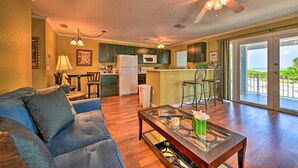 A living room with a blue sofa, a glass coffee table, and a kitchen area with a refrigerator, microwave, and bar stools. - Bright Coastal Condo: Walk to Lido Key Beach! (Sarasota)