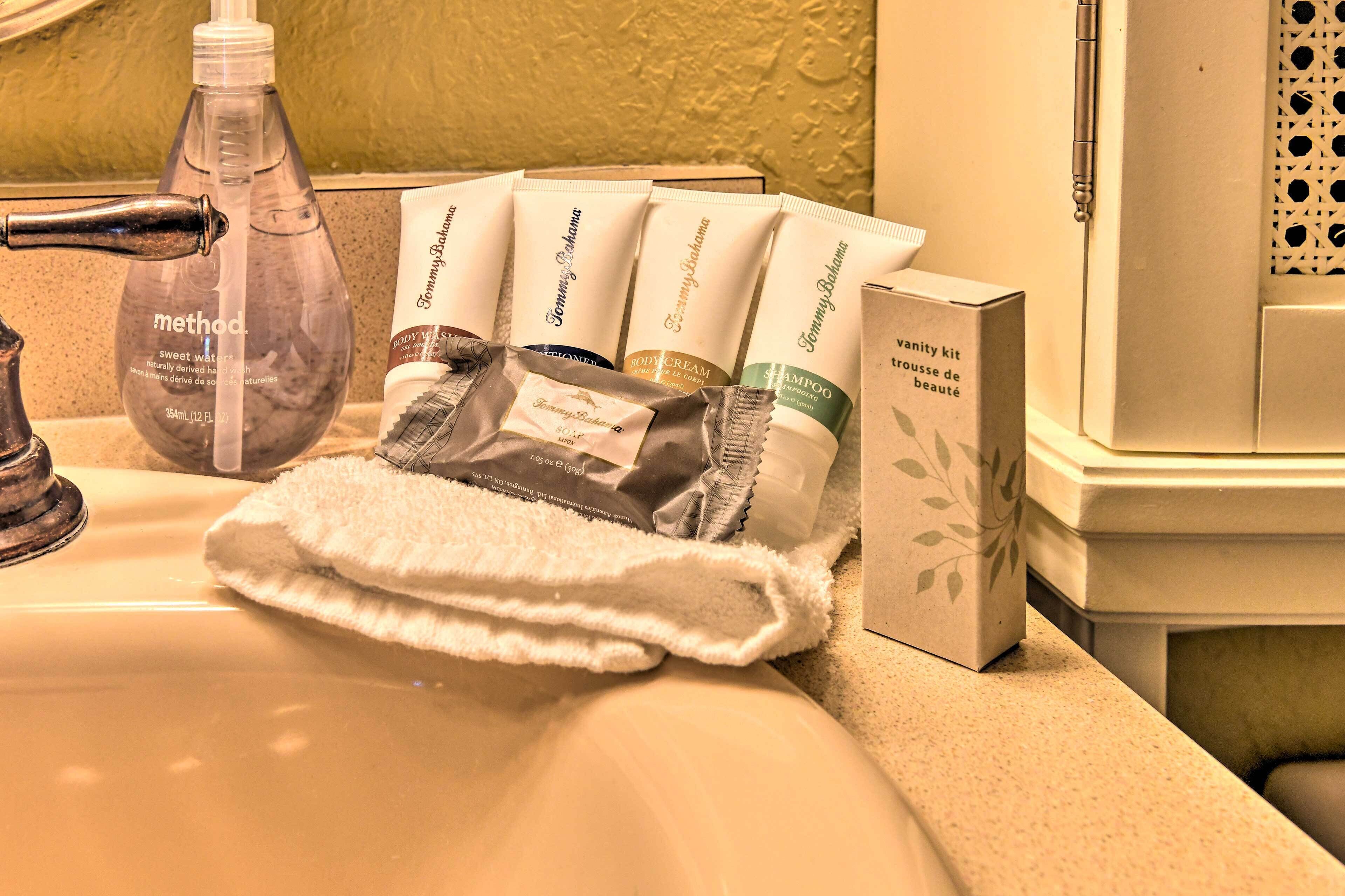 A bathroom sink with toiletries including a bottle of Method body wash, several Tommy Bahama body care products, and a vanity kit box.