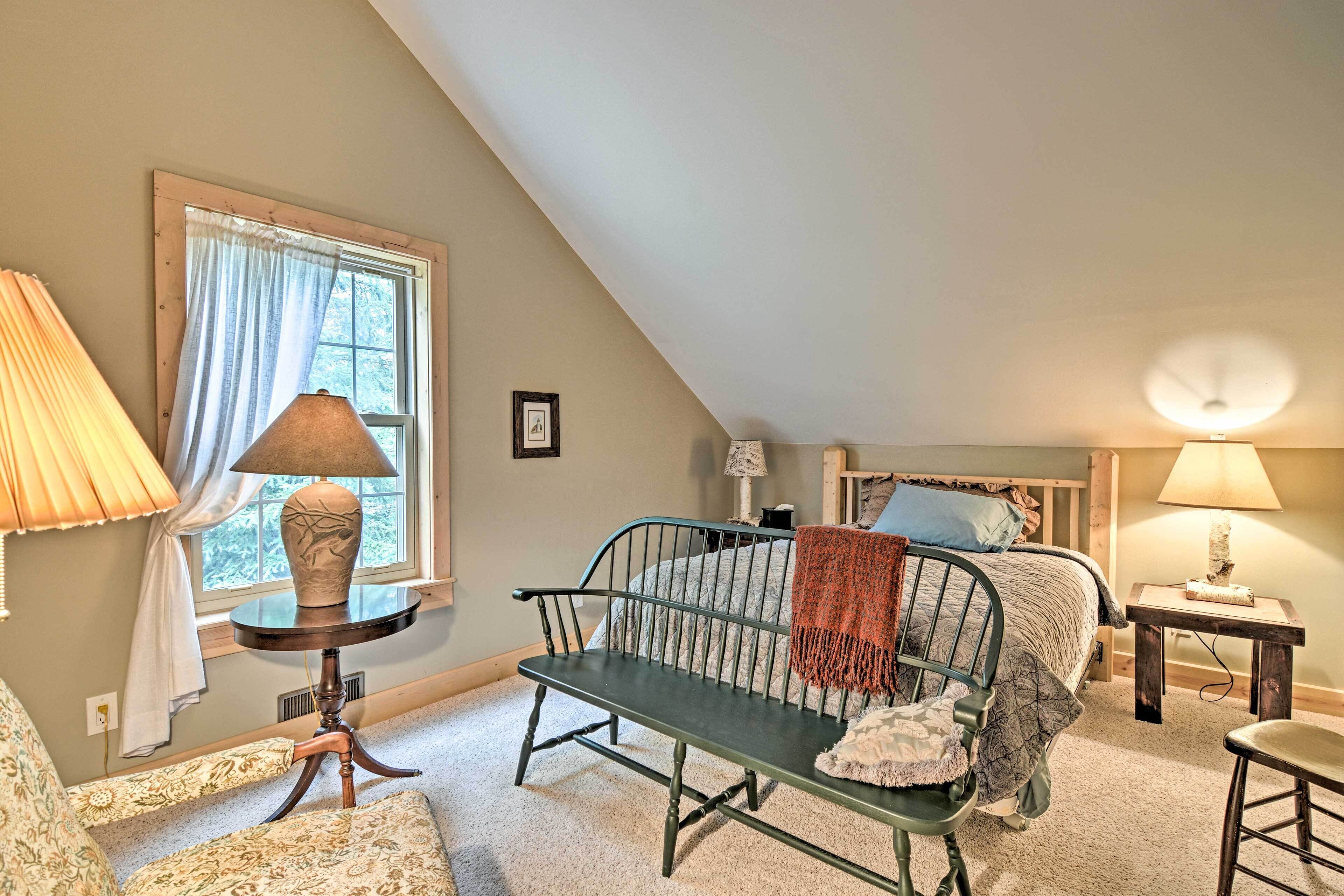 A cozy bedroom with a sloped ceiling, a bed with a blue headboard, a bench, a chair, and two lamps.