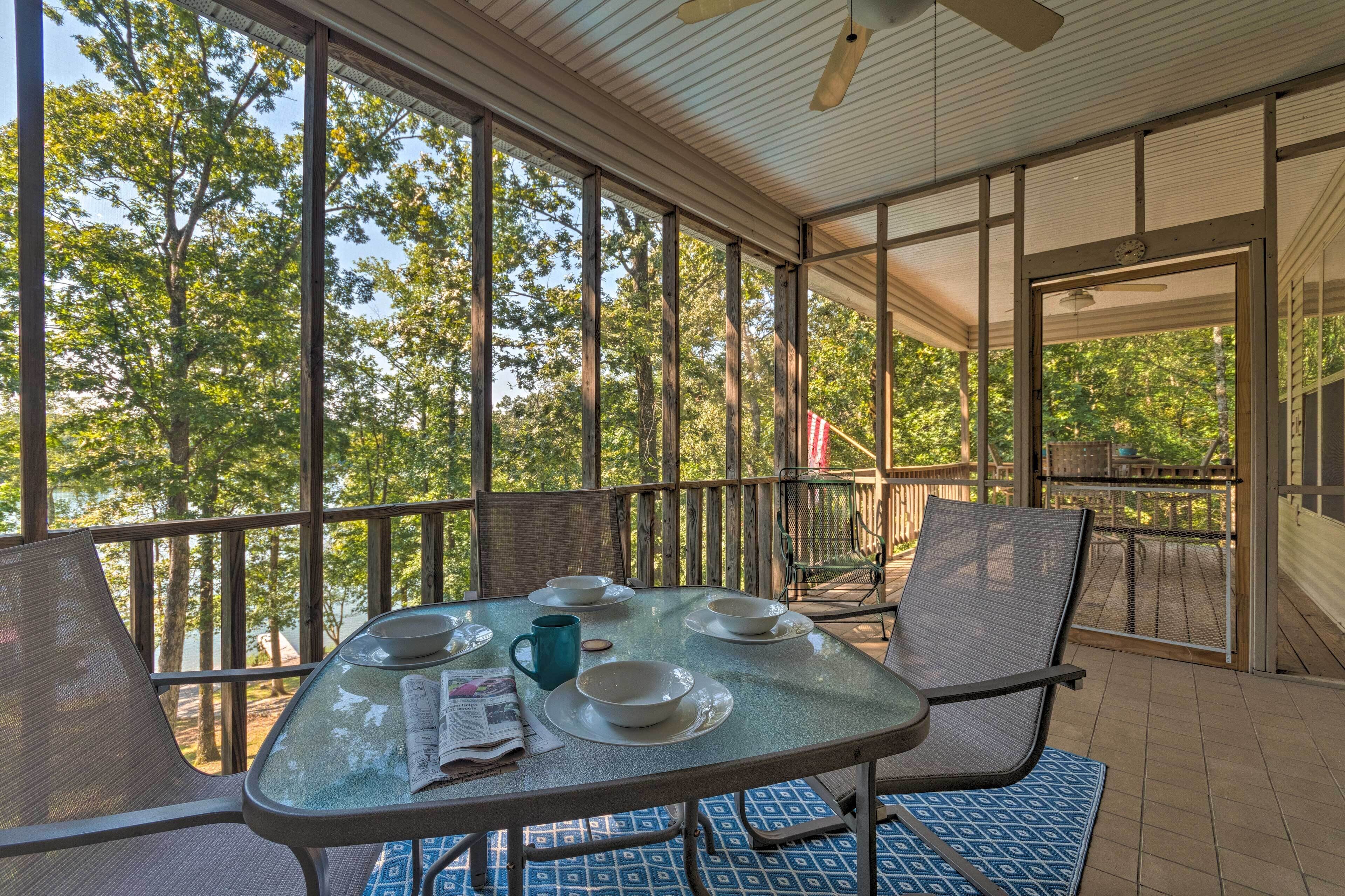 A covered outdoor dining area with a table set for four, surrounded by chairs, overlooking a wooded area.