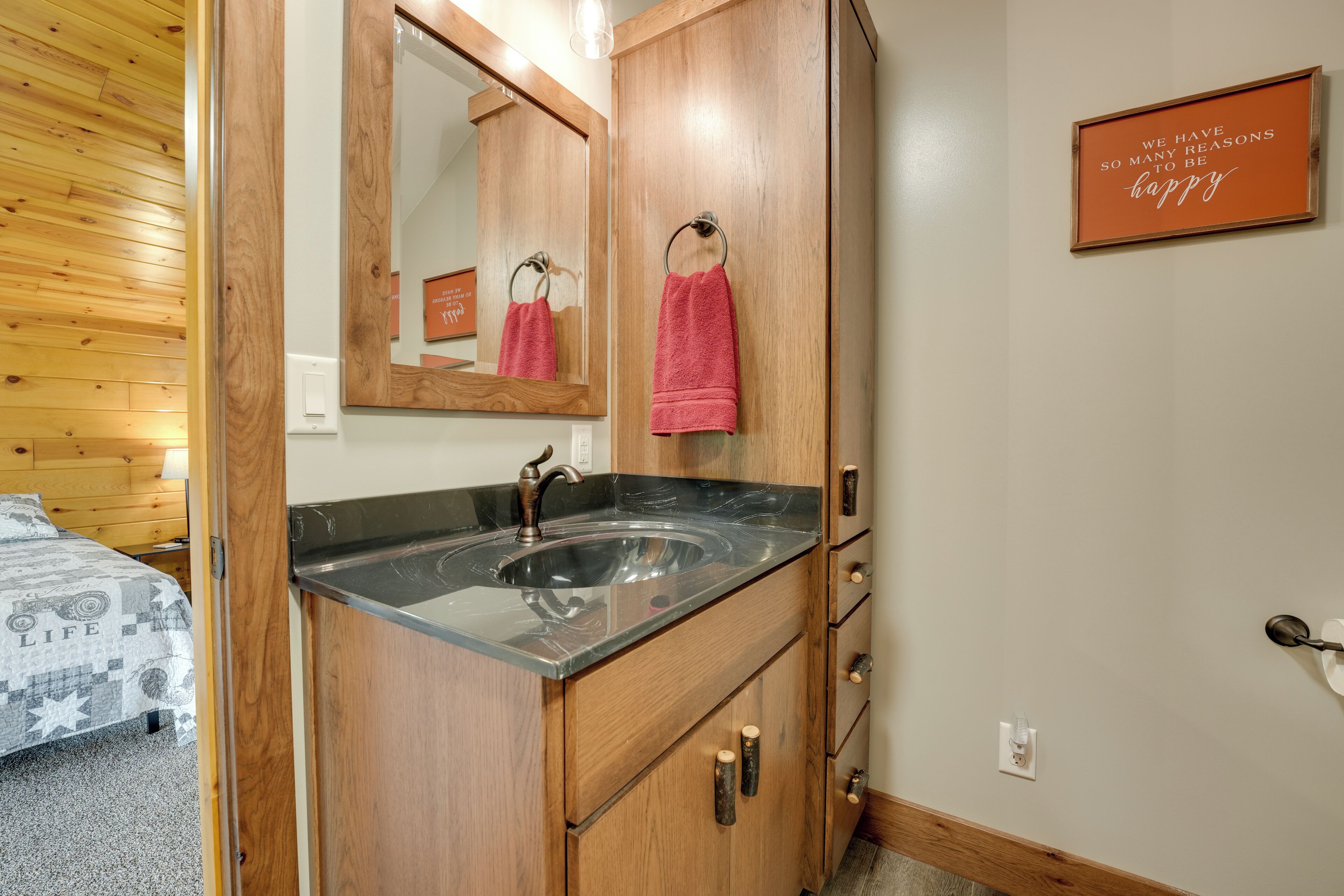 A bathroom with a wooden vanity, a black sink, and a mirror. There is a towel rack with a pink towel hanging on it. A sign on the wall reads "We have so many reasons to be happy."