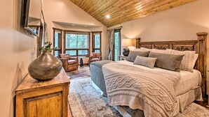 A bedroom with a wooden ceiling, a large bed with multiple pillows, a wooden nightstand, a vase on a dresser, and a window with a view of trees.