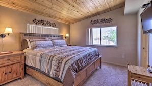 A bedroom with a wooden ceiling, a large bed with patterned bedding, two bedside lamps, a window with a view, and a TV on the wall. - Rustic Bullhead City Retreat w/ Porch & Views (Bullhead City)