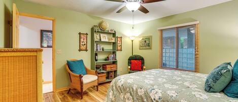 Quarto com cama, parede verde, estante, cadeira de vime, janela com persianas e ventilador de teto.