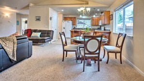 A spacious living room with a black leather sofa, a dining area with wooden chairs and a table, and a kitchen with wooden cabinets and a window.
