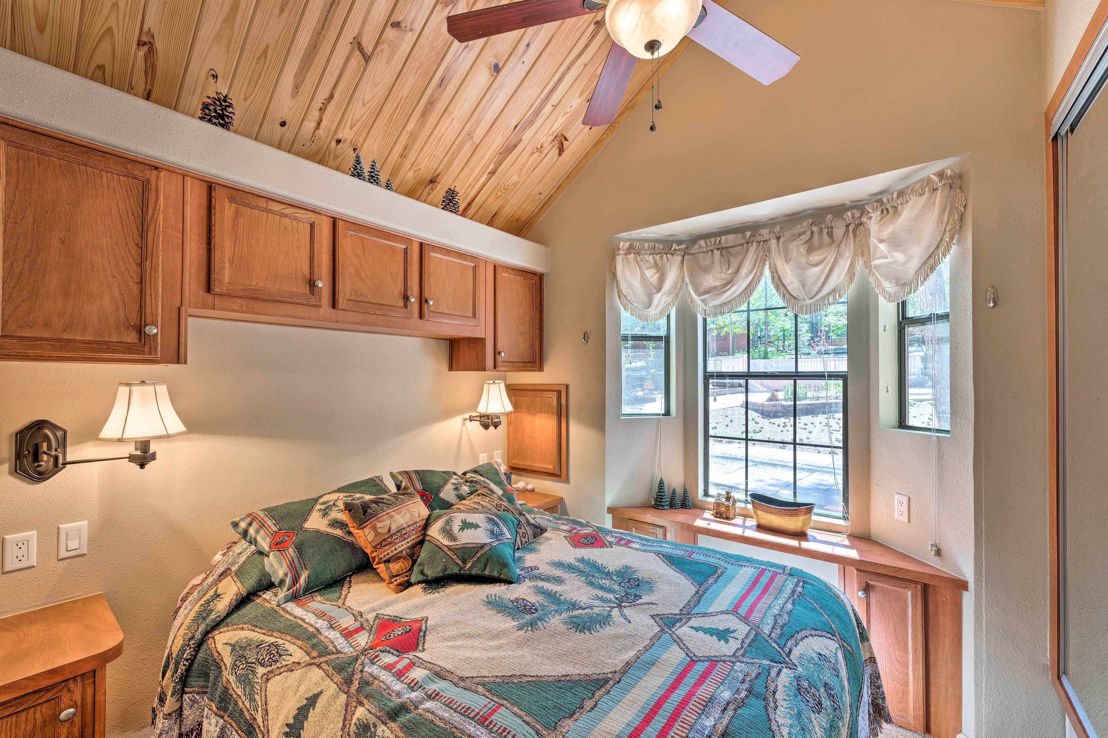 A bedroom with wooden ceiling, a bed with a quilt, two bedside lamps, a window with curtains, and a small table with a bowl.
