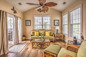 Un salon comprenant un canapé, une table basse et des fauteuils. On y trouve également un ventilateur de plafond, une fenêtre avec des stores et une porte avec des rideaux.