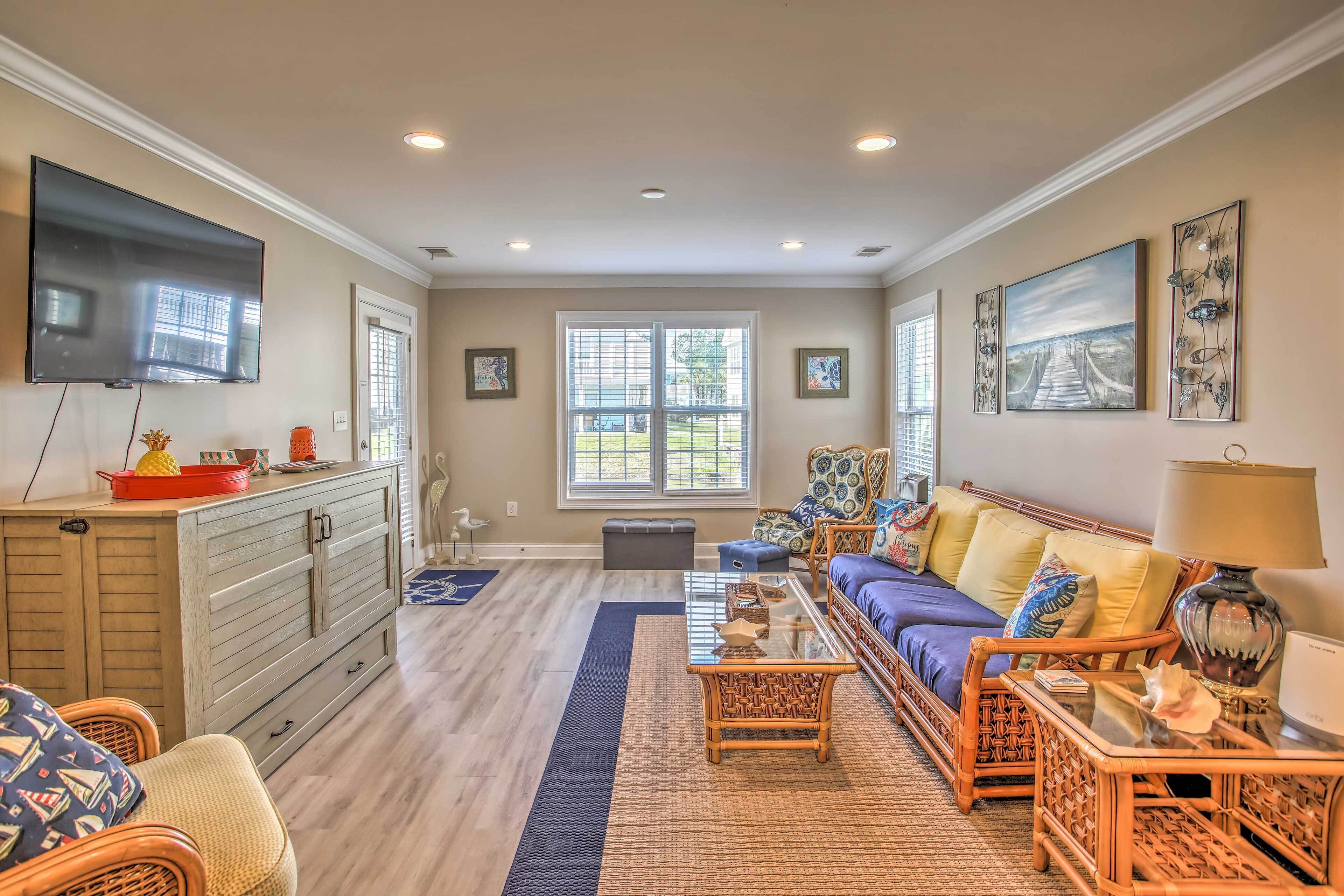 A spacious living room with a large flat-screen TV, a sofa set, wicker furniture, and artwork on the walls.