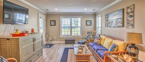 A spacious living room with a large flat-screen TV, a sofa set, wicker furniture, and artwork on the walls.