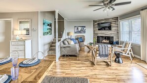 A spacious living room with a fireplace, a dining area with a wooden table, and a staircase leading to another level. - Waterfront Home w/ Decks, Privacy & Boat Slip (Supply)