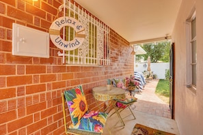 A patio area with a brick wall, a decorative sign, a chair with a floral cushion, a small round table, and a view of the backyard.