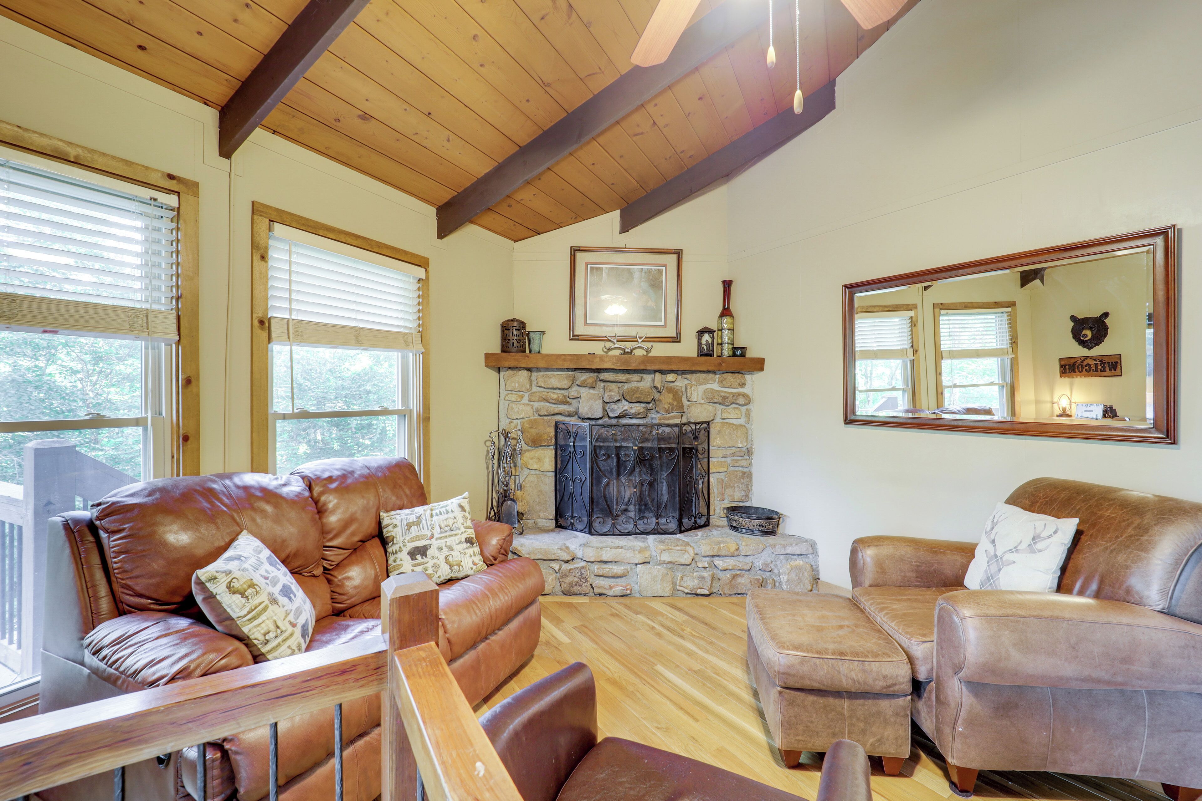 A cozy living room with leather sofas, a stone fireplace, wooden beams, and large windows.