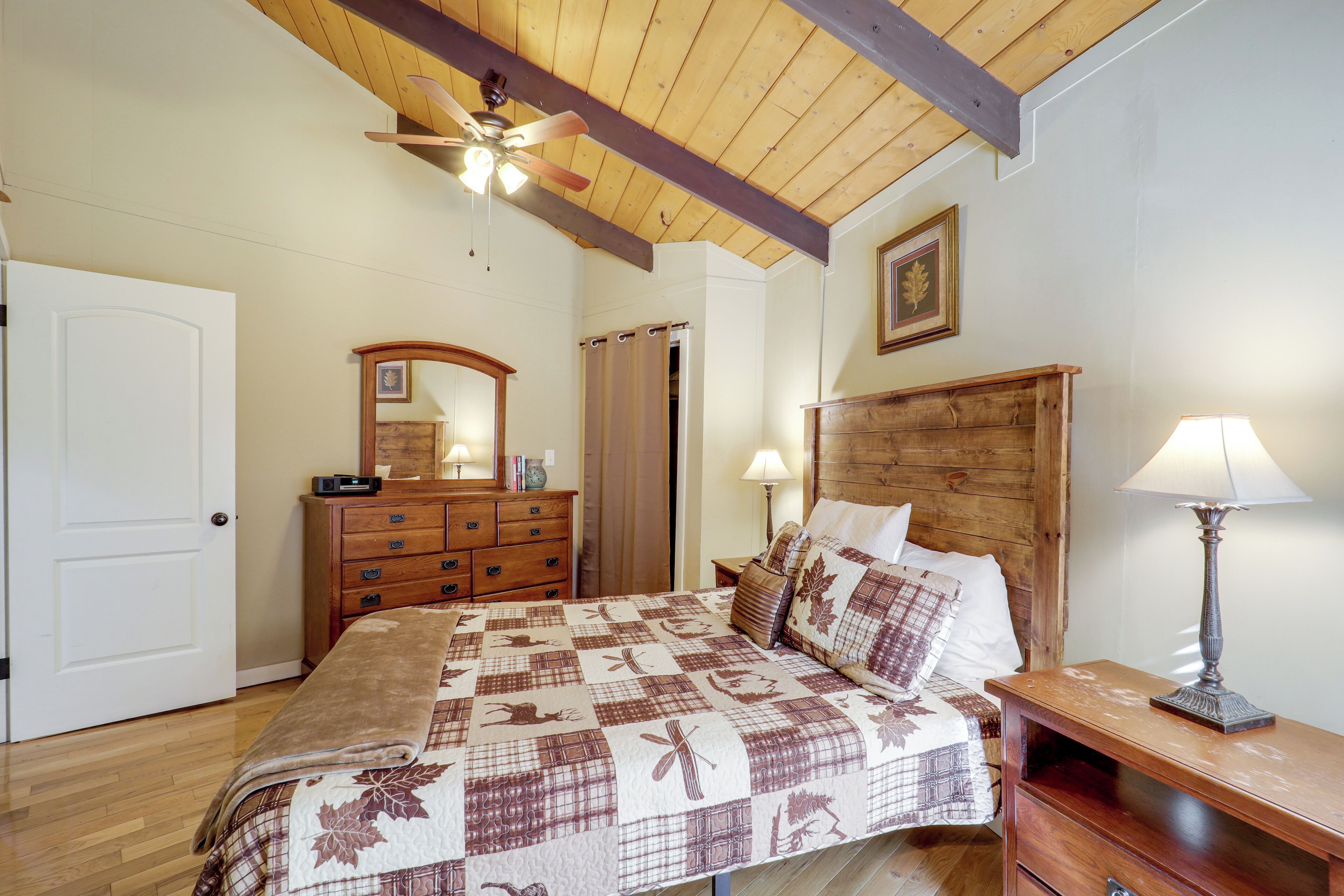 Een slaapkamer met een houten plafond, een bed met een quilt met bloemenprint, een dressoir, een nachtkastje met een lamp en een deur.