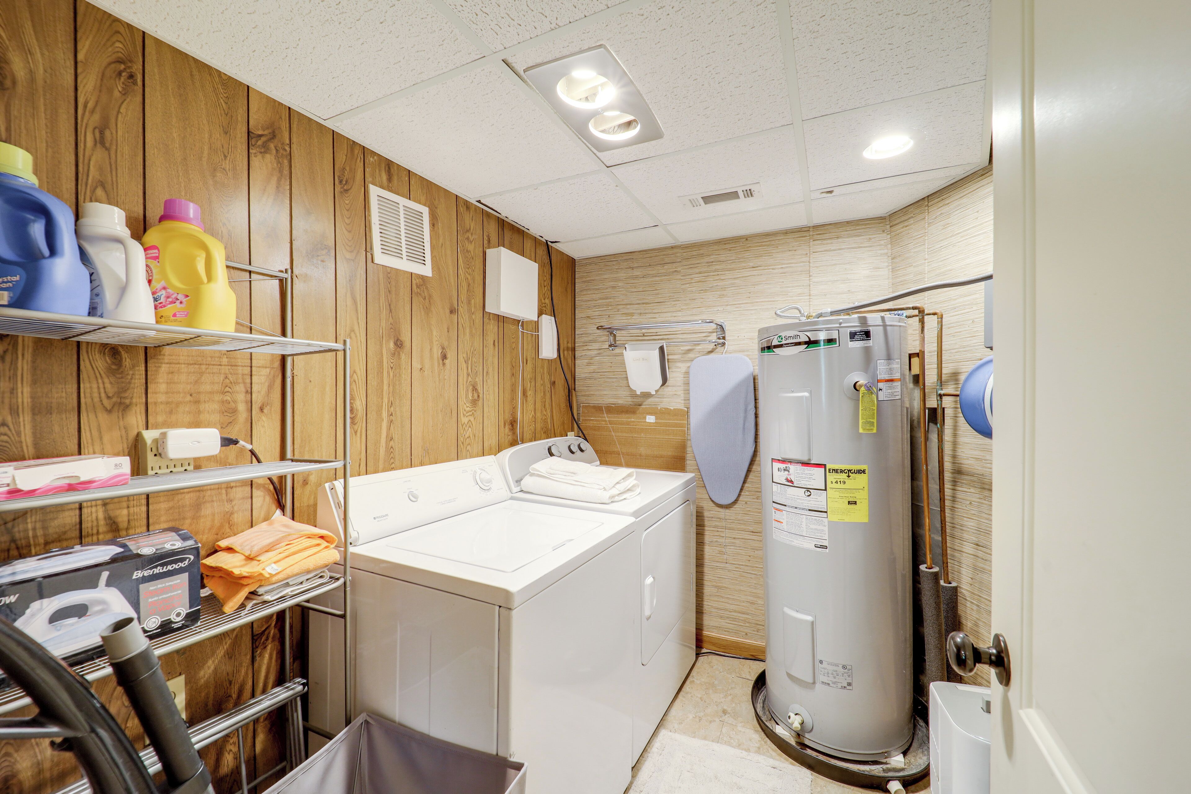 A laundry room with a front-loading washer and dryer, a shelf with cleaning supplies, and a water heater.