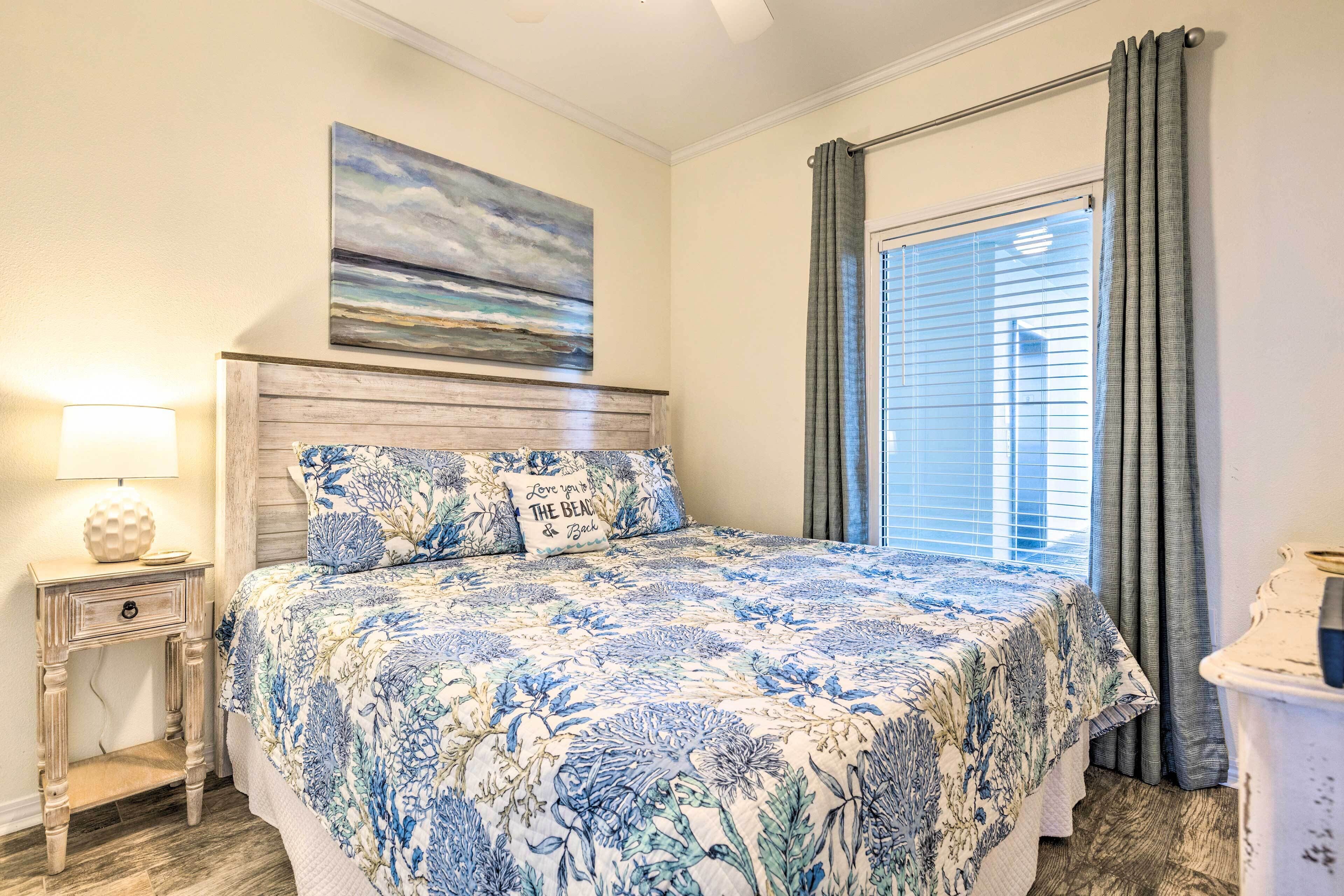 A bedroom with a floral bedspread, a nightstand with a lamp, a painting above the bed, and a window with curtains.
