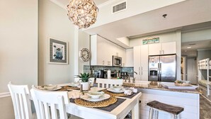 A modern kitchen with white cabinetry, a central island, and a dining area with white chairs and a wooden bench. - Ocean Views & Pool Access: Orange Beach Condo! (Orange Beach)
