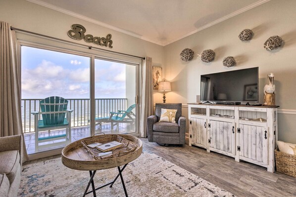 A living room with a large sliding glass door leading to a balcony with chairs, a television on a wooden cabinet, and a decorative wall piece. - Ocean Views & Pool Access: Orange Beach Condo! (Orange Beach)