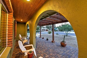 A covered patio area with brick flooring, white chairs, and potted plants. - Lovely Phoenix Home w/ Expansive Patio & Fire Pit! (Phoenix)