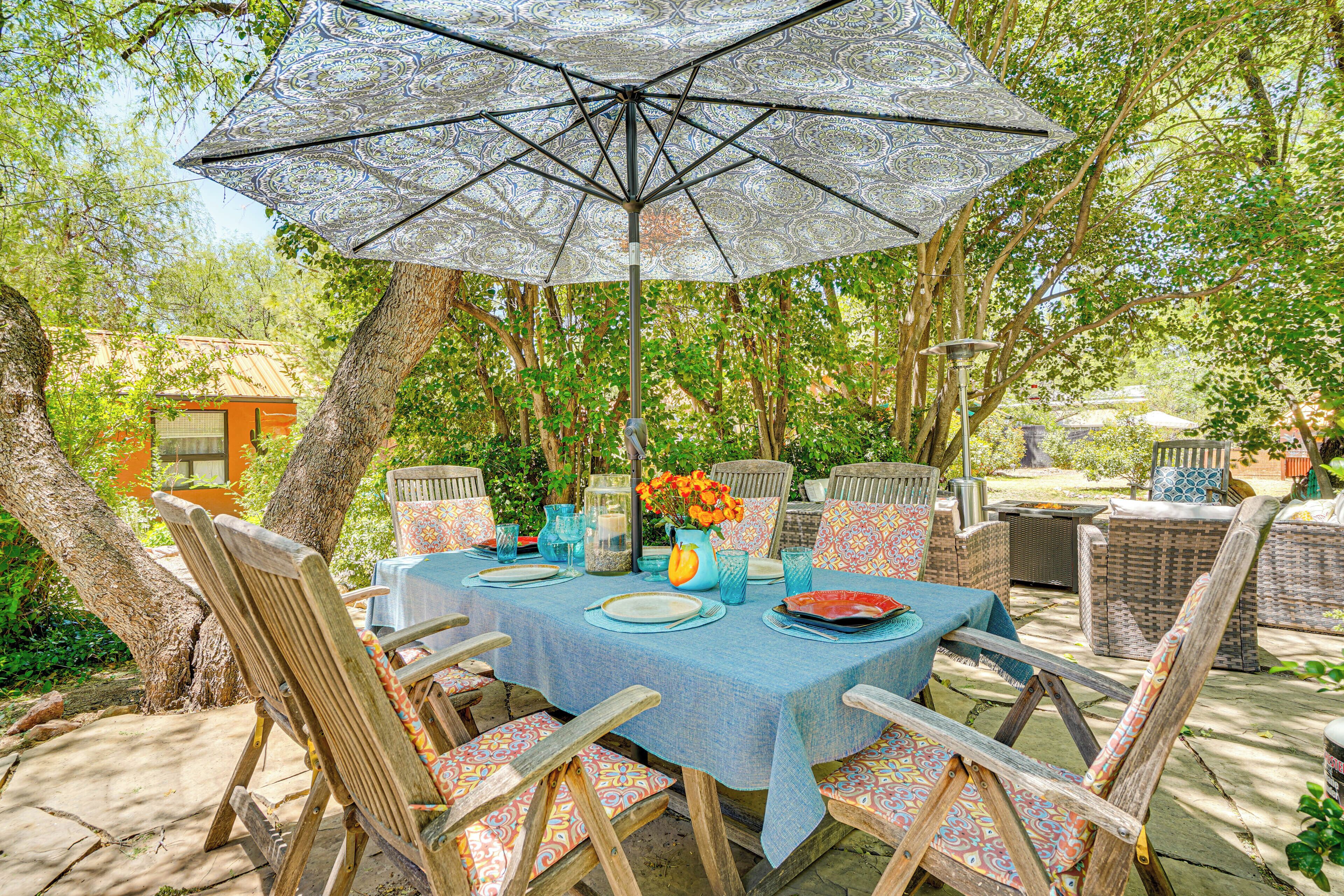 An outdoor dining area with a table set for six, surrounded by wicker chairs, under a patterned umbrella.
