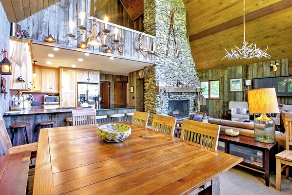 A rustic cabin interior with a wooden dining table, stone fireplace, and a stone chimney.