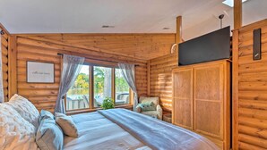 Un dormitorio de cabaña de troncos con una cama grande, un televisor en la pared y una ventana con cortinas.