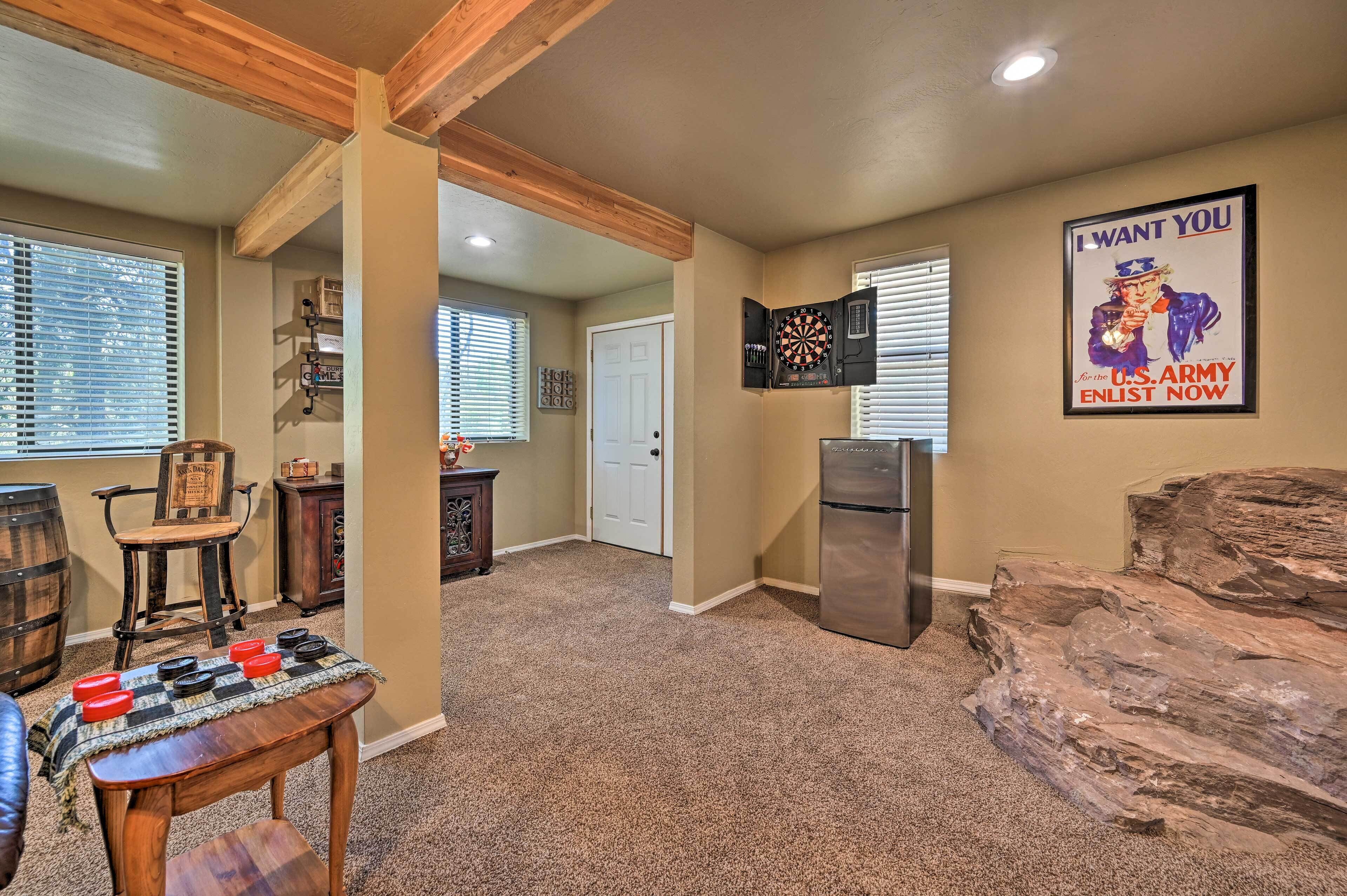 A spacious living area with a dartboard, a wooden chair, a table with darts, a refrigerator, and a rock feature.