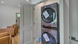A modern laundry room with stacked washers and dryers, a door leading to a living area with sofas, and a bottle of detergent on the floor. - Rural Family Retreat < 3 Mi to Fishing & Boating! (Higden)
