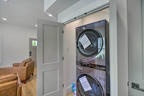 Une buanderie moderne avec des lave-linge et des sèche-linge superposés, une porte donnant sur un espace de vie avec des canapés, et une bouteille de lessive posée sur le sol.