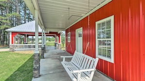 A covered porch with a white bench, a red building with white-framed windows, and a stone structure in the background. - Rural Family Retreat < 3 Mi to Fishing & Boating! (Higden)