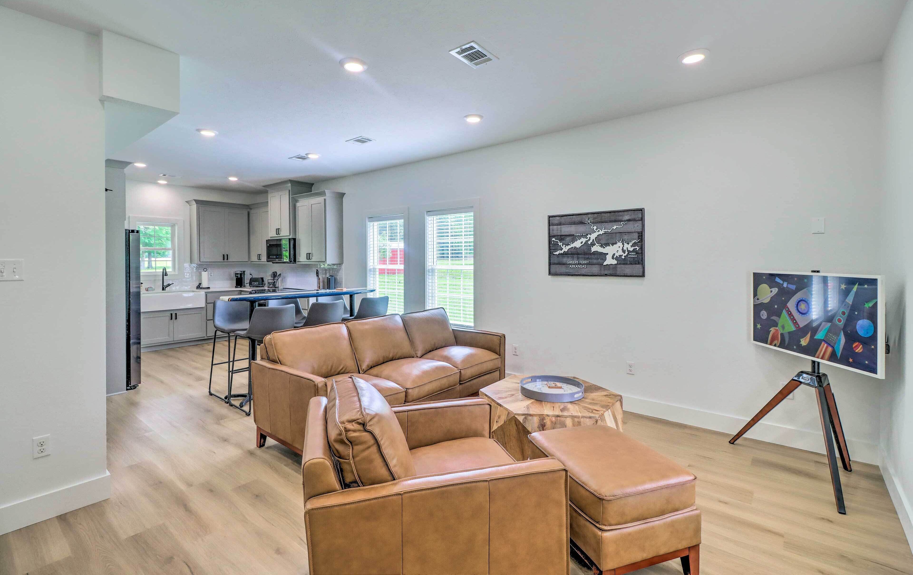 A spacious living room with a leather sofa, a dining area with a table and chairs, a kitchen in the background, and artwork on the walls.