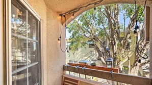 A balcony with a wooden bench, a window with a view of trees and buildings, and string lights.