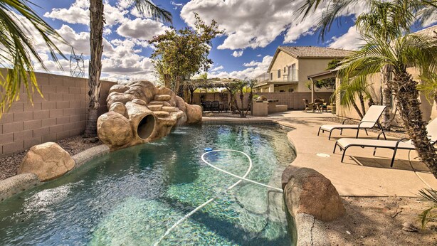 Une piscine entourée de rochers et de palmiers, avec des chaises longues et une maison à proximité.