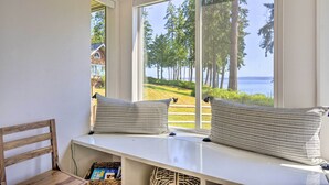 A room with a large window overlooking a forest and a body of water. There is a bench with two pillows and a wooden chair. - Hike, Bike & Explore: Oceanfront Kingston Getaway (Kingston)