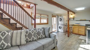 A living room with a gray sofa, patterned pillows, a wooden staircase, and a kitchen area with a washing machine. - Cozy 'hillside Hideaway' in Wintergreen Resort! (Roseland)