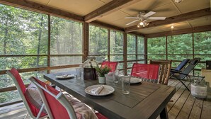 A wooden deck with a table set for dining, surrounded by chairs, and large windows offering a view of the forest. - Mountain Town Adventures Home w/ Fire Pit! (Fort Valley)