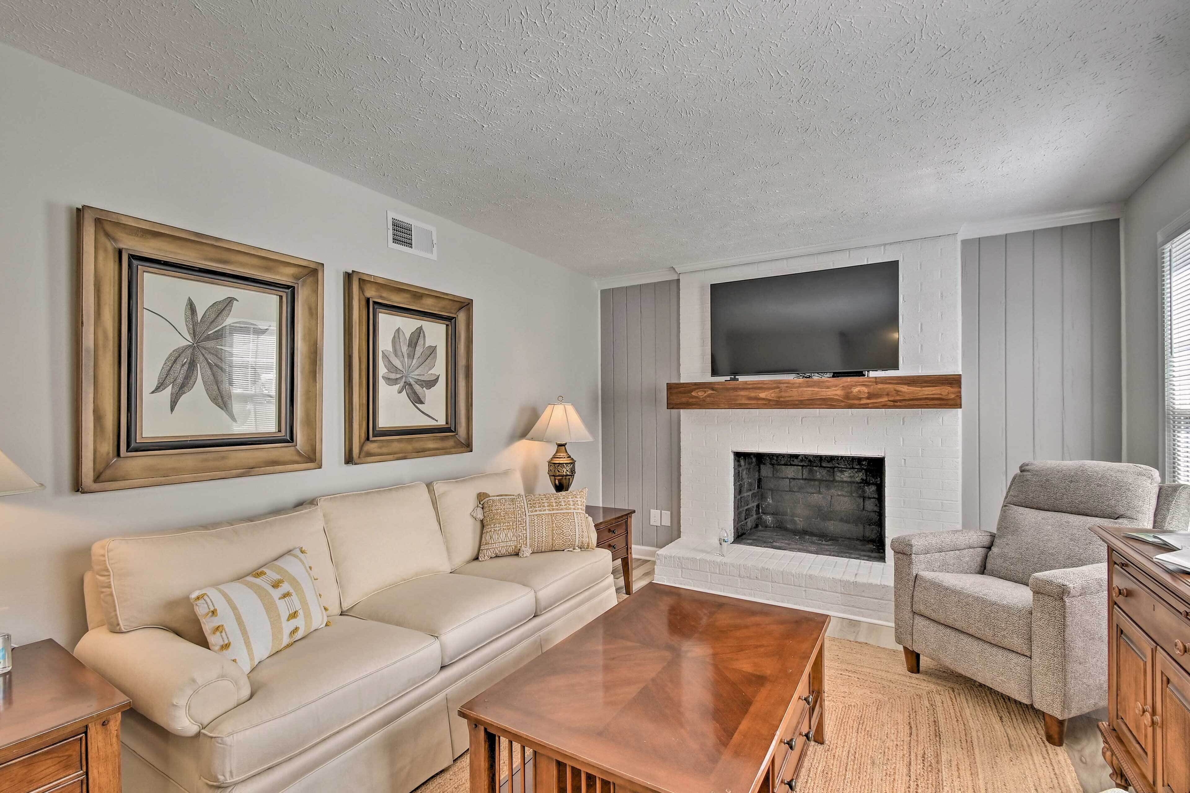 A living room with a beige sofa, a wooden coffee table, a grey armchair, a fireplace, and two framed floral artworks on the wall.
