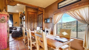 A rustic dining area with a wooden table and chairs, a view of the desert through a large window, and a cozy living space in the background. - Mtn-view Adobe Home w/ Large Deck Near Prescott! (Mayer)