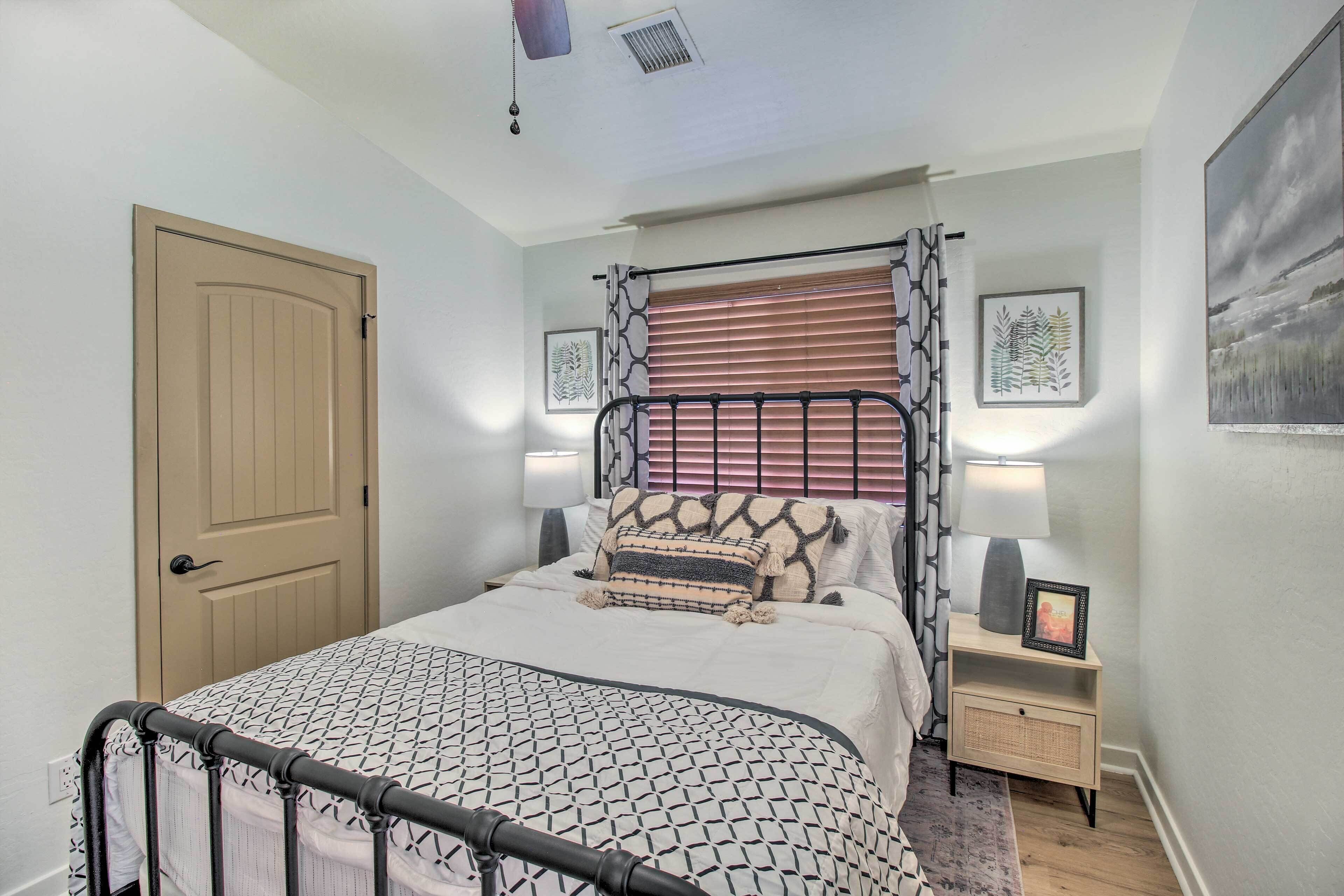 A bedroom with a bed, a nightstand, a lamp, a window with blinds, a door, and a framed picture on the wall.